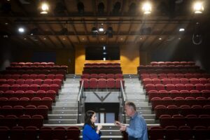 Dos personas conversando en un teatro con asientos rojos vacíos.