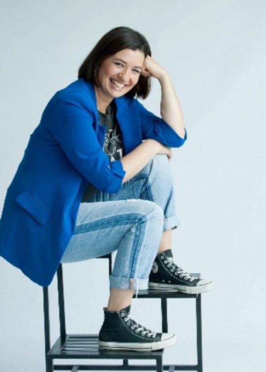 Mujer sonriente sentada en una silla, vestida con chaqueta azul y jeans.