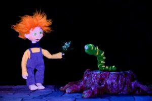Títere con cabello naranja interactuando con un gusano en un espectáculo infantil.