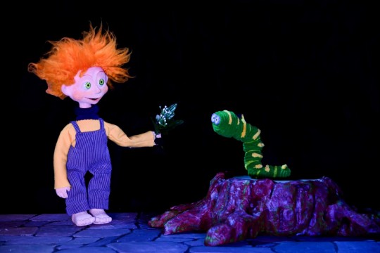 Títere con cabello naranja interactuando con un gusano en un espectáculo infantil.