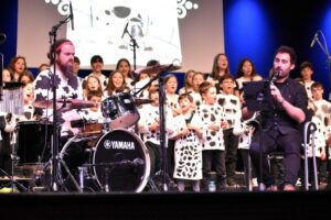 Músicos en un escenario con un coro de niños de fondo