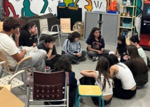 Grupo de jóvenes en ensayo teatral, sentados en círculo en un espacio comunitario.
