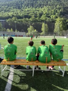 Niños sentados en un banco observando un partido de fútbol en un campo verde