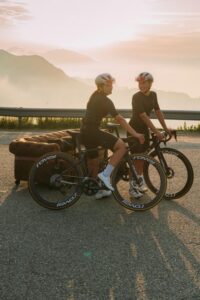 Dos ciclistas en una carretera con un sofá al fondo durante el amanecer.