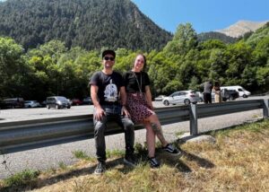 Dos personas sentadas en una barandilla con paisaje montañoso detrás.