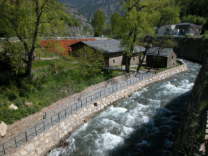 Conjunto de edificios de piedra junto a un río, con pasarela y árboles