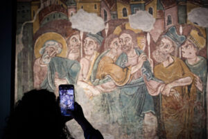 Una persona fotografía con su teléfono una pintura mural medieval con varias figuras.