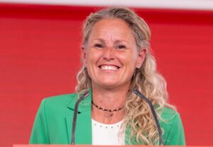 Mujer sonriente con chaqueta verde frente a dos micrófonos y fondo rojo