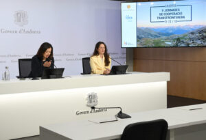 Dos mujeres sentadas ante una mesa, con una pantalla que anuncia las jornadas de cooperación transfronteriza.